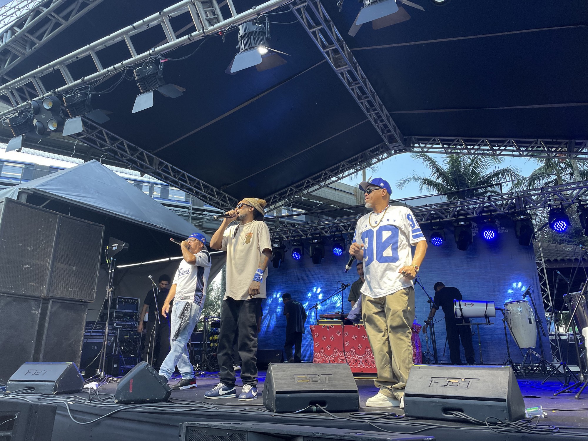 #PraTodosVerem: Doctor MC's se apresentam no palco. Eles usam bonés e roupas largas, um deles veste camiseta branca de time de futebol americano com o número 90. Todos estão com microfones nas mãos, interagindo com o público. O palco é iluminado por luzes azuis.