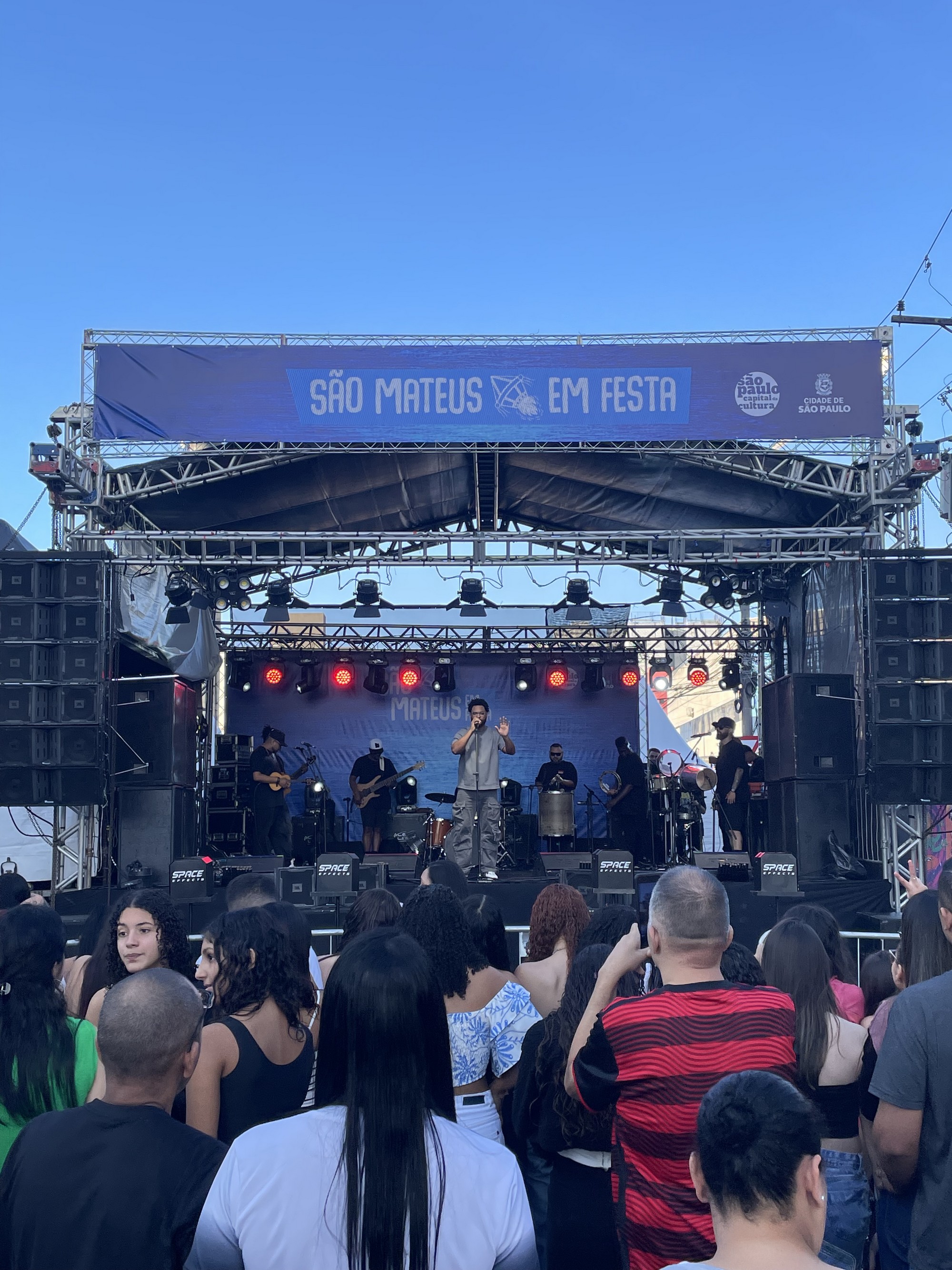 #PraTodosVerem: Foto tirada na visão do público, que assiste ao cantor Julio Sereno no palco. O banner no alto diz “São Mateus em Festa”. No palco, o artista veste camiseta cinza e calça escura, cantando com as mãos erguidas. Músicos ao fundo tocam instrumentos como cavaquinho e percussão, enquanto refletores iluminam o espaço.