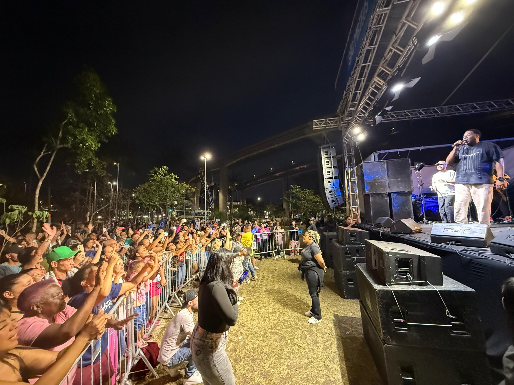 #PraTodosVerem: O cantor Netinho de Paula veste camiseta preta e calça clara, segurando o microfone enquanto se apresenta no palco à noite. Ao lado dele, outros músicos acompanham a performance. Na frente do palco, uma grande multidão atrás das grades ergue os braços e canta junto, muitos com celulares levantados para gravar. No canto inferior direito, uma mulher de camiseta preta e calça jeans observa o show próxima às caixas de som. Árvores iluminadas e postes de luz compõem o fundo da cena.