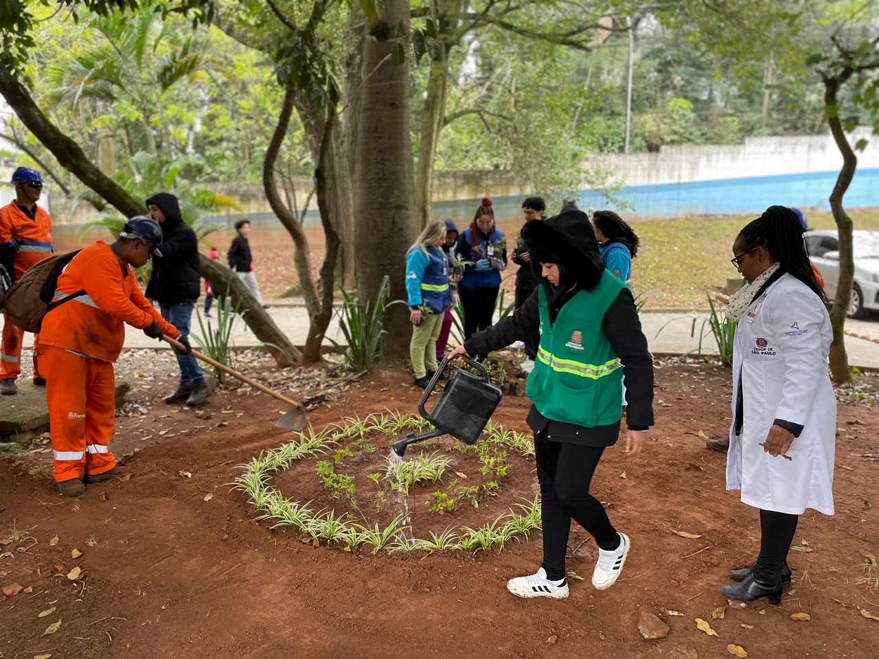 #PraTodosVerem: Espaço aberto, dois homens de uniforme laranja, um deles trabalhando nas mudas de plantas na terra. Uma mulher de uniforme verde rega as plantas e as pessoas de fundo conversam.