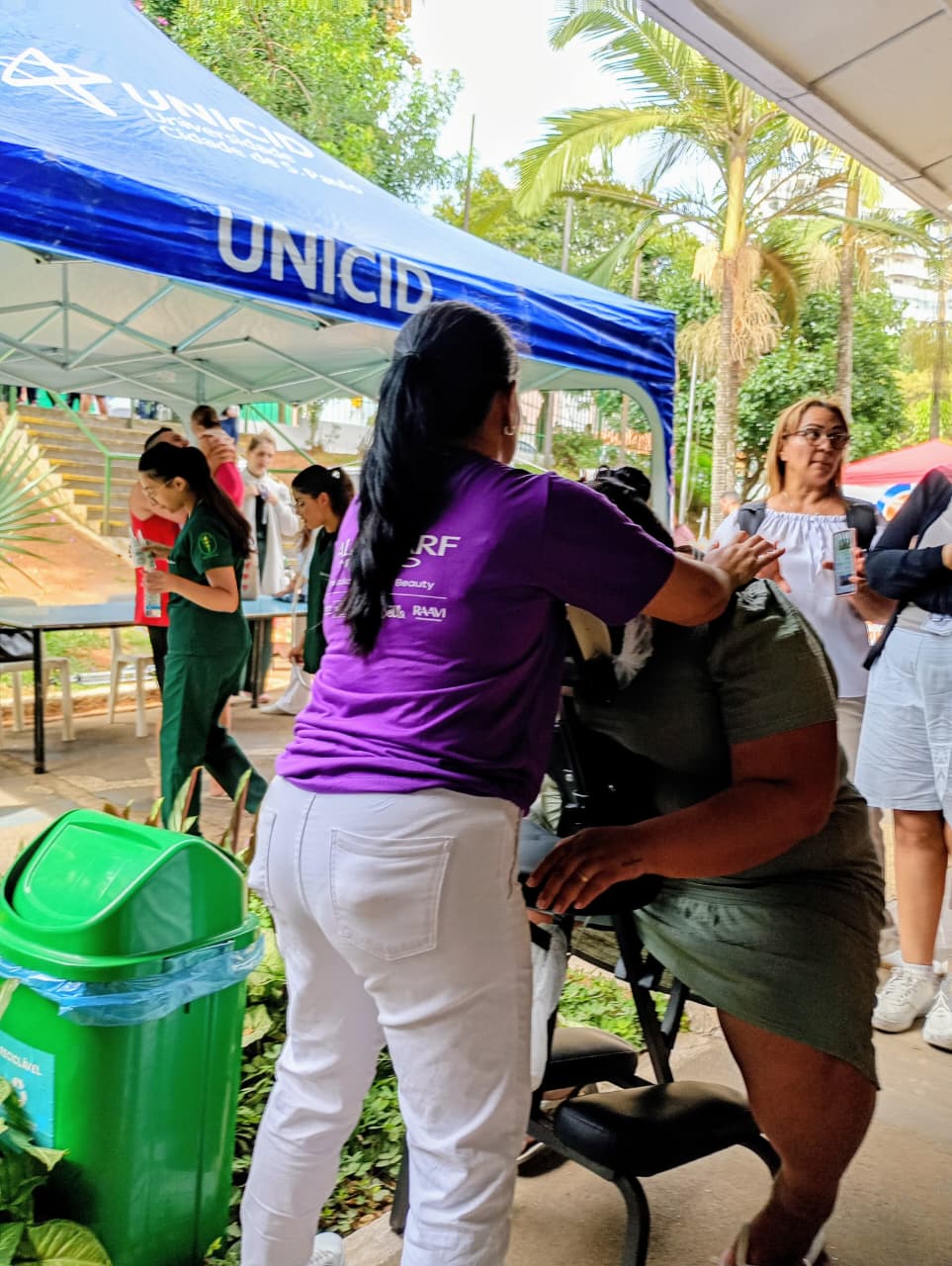 Uma pessoa está sentada recebendo massagem em uma ação de Dia das Mães da Subprefeitura de São Miguel Paulista. O atendimento acontece em um espaço aberto, com outras pessoas ao redor e uma tenda azul ao fundo.