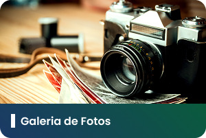 Pra Todos Verem: Card com fotografia de uma câmera fotográfica posicionada sobre uma superfície de madeira. Ao lado da câmera, há fotografias impressas parcialmente espalhadas, sugerindo o registro e a preservação de imagens. A composição utiliza tons quentes e foco nos equipamentos e materiais relacionados à fotografia. Na faixa inferior em degradê azul e verde, está o texto: “Galeria de Fotos”. A arte representa um espaço destinado à visualização e ao compartilhamento de registros fotográficos.