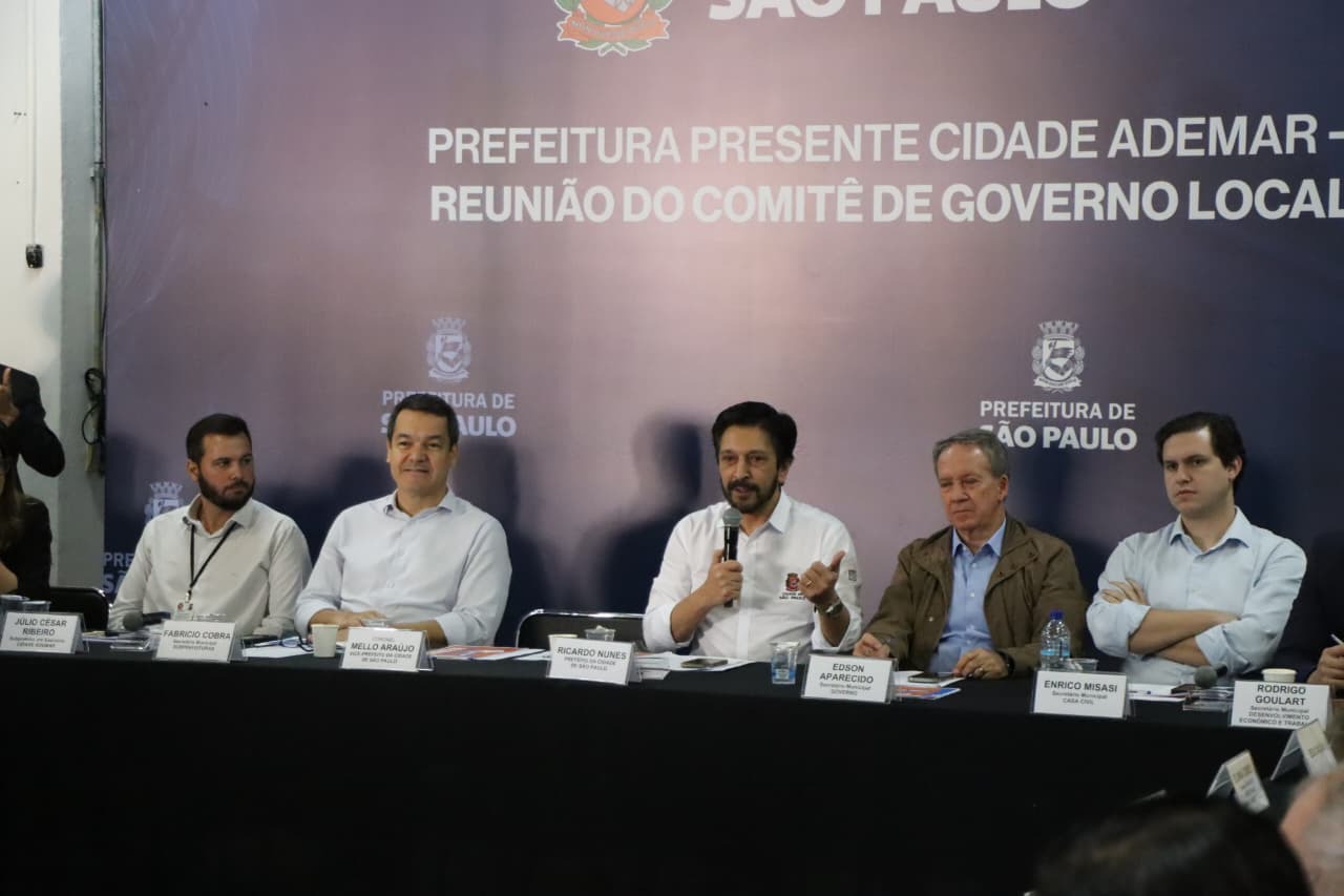 Uma fotografia em primeiro plano de cinco homens sentados lado a lado em uma mesa, durante uma reunião. Eles usam camisas de manga comprida e estão voltados para a frente. O homem ao centro, com barba, segura um microfone e está falando. As mesas têm toalhas pretas. Em frente a eles, há placas de identificação com seus nomes e cargos. Da esquerda para a direita, é possível ler "JULIO CÉSAR BREJO", "FABRICIO CORNA", "MELLO ARAUJO", "RICARDO NUNES", "EDSON APARECIDO", "ENRICO MISASI" e "RODRIGO GOULART". Ao fundo, um painel azul com o brasão da cidade de São Paulo e o texto "PREFEITURA PRESENTE CIDADE ADEMAR - REUNIÃO DO COMITÊ DE GOVERNO LOCAL".