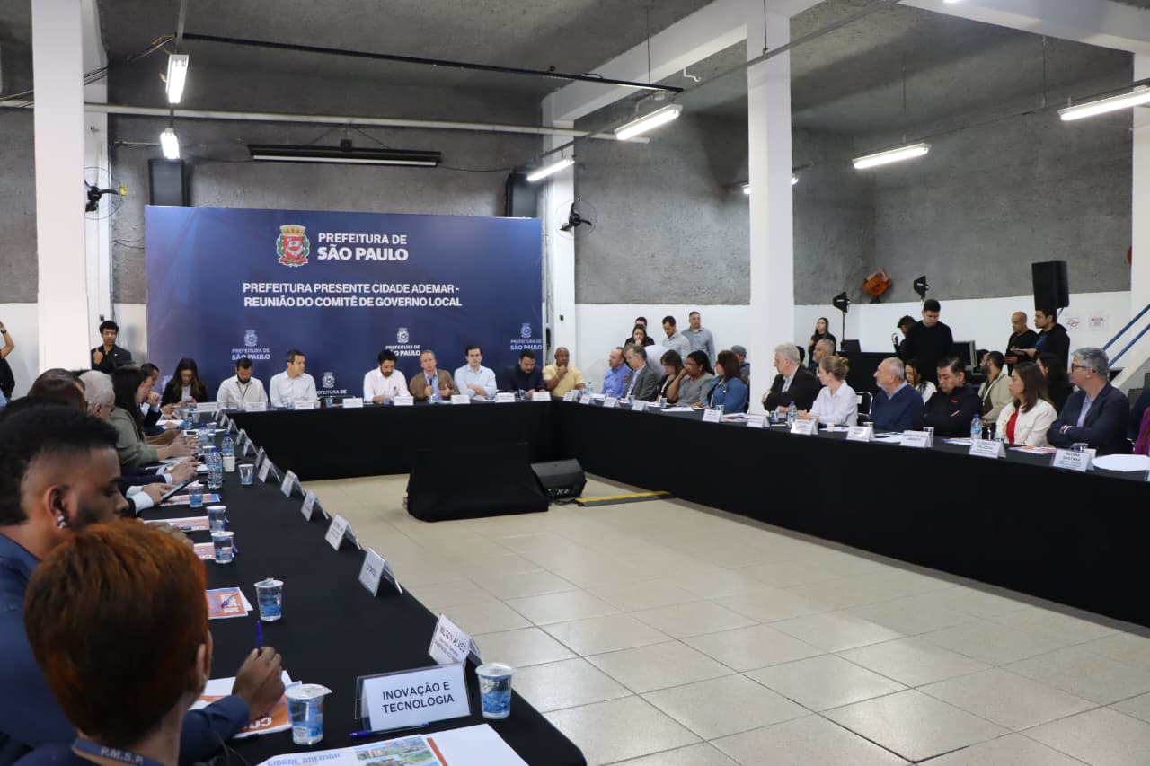 Uma reunião do Comitê de Governo Local da Prefeitura de São Paulo. A imagem mostra duas longas mesas formando um "L", onde estão sentados diversos participantes, a maioria de meia-idade e usando roupas sociais. Eles observam e conversam entre si, com olhares voltados para a frente da sala. No centro, ao fundo, um grande painel azul com o brasão da cidade e o texto "PREFEITURA DE SÃO PAULO" em destaque. Abaixo, em letras brancas menores, está escrito "PREFEITURA PRESENTE CIDADE ADEMAR - REUNIÃO DO COMITÊ DE GOVERNO LOCAL". Várias pessoas assistem de pé nos cantos da sala, e um homem de camisa escura fala ao microfone. As mesas estão cobertas com toalhas pretas, e há garrafas d'água, copos e pastas sobre elas. Há uma placa branca à frente que diz "INOVAÇÃO E TECNOLOGIA".