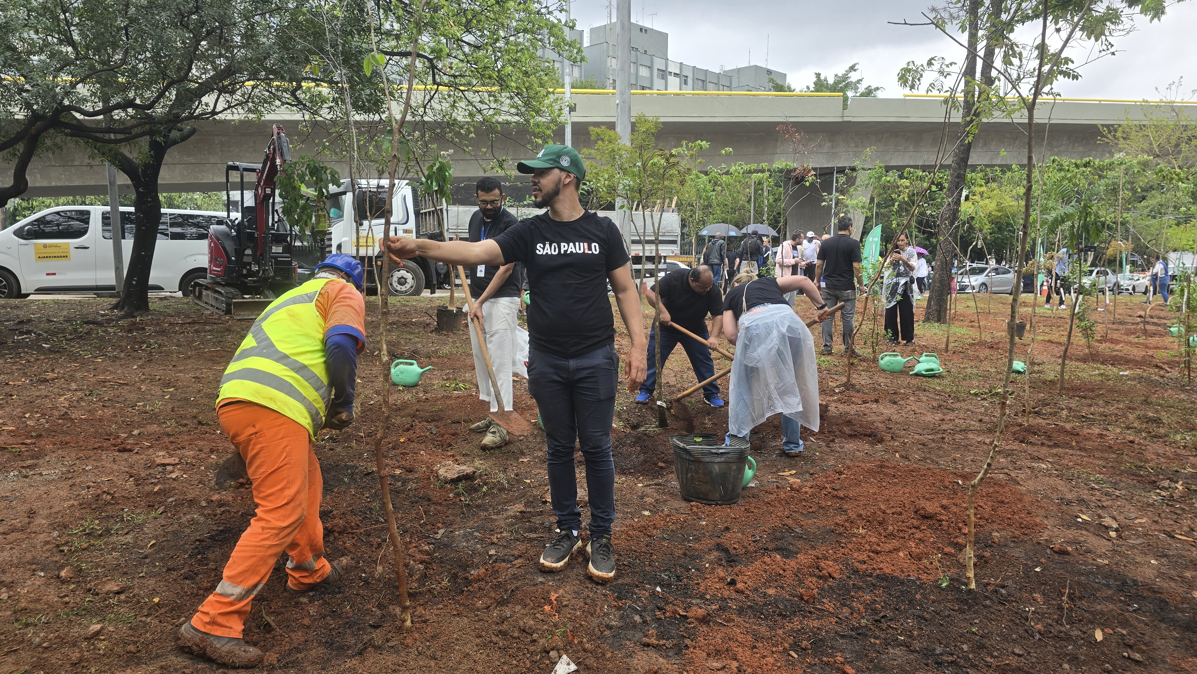 Fotografia em um local de plantio de árvores, com solo de terra vermelha exposta. Várias pessoas estão participando do plantio, em um esforço comunitário ou de equipe.  Em primeiro plano à esquerda, um trabalhador da limpeza ou jardinagem está de costas, curvado, vestindo uniforme laranja e um colete refletor amarelo e azul, auxiliando no plantio de uma muda de árvore.  No centro, um homem de camiseta preta com a inscrição "SÃO PAULO" em branco, boné verde e calça jeans escura, está de pé e gesticulando em direção à muda que está sendo plantada.  Ao redor, outras pessoas, algumas vestindo roupas casuais escuras e outras vestindo aventais de proteção, estão engajadas em cavar e regar as mudas (pequenos regadores verdes estão espalhados pelo chão).  Ao fundo, sob um viaduto de concreto com uma proteção lateral amarela, há veículos estacionados ou em movimento e um grupo maior de pessoas, indicando um evento de plantio em área urbana. O dia está nublado.