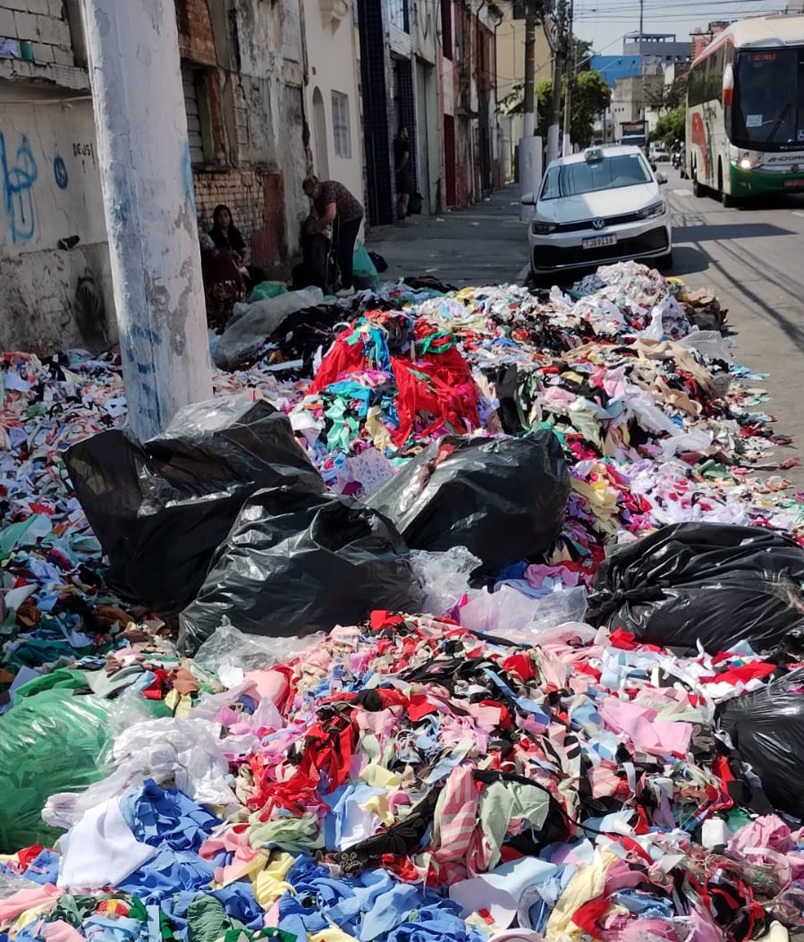  Pracegover: Fotografia de uma cena urbana mostrando um grande volume de resíduos têxteis ou retalhos de tecido espalhados pela calçada e parte da sarjeta, em um dia ensolarado.  Em primeiro plano e no centro, uma enorme pilha de retalhos de diversas cores (principalmente vermelho, rosa, azul, branco e preto) se estende ao longo da calçada. Vários sacos plásticos de lixo pretos e verdes, cheios, estão espalhados no meio dos retalhos.  Ao fundo, a rua é ladeada por construções baixas e antigas. No lado esquerdo, sobre a calçada, um grupo de pessoas está próximo a uma parede pichada, aparentando estar interagindo com os resíduos ou o local.  Na rua, à direita, há um carro de cor clara (aparentemente um táxi) estacionado ou em movimento lento, e um ônibus de transporte público de cor verde, branco e vermelho está se afastando no fundo. Um poste de luz está em primeiro plano, à esquerda.
