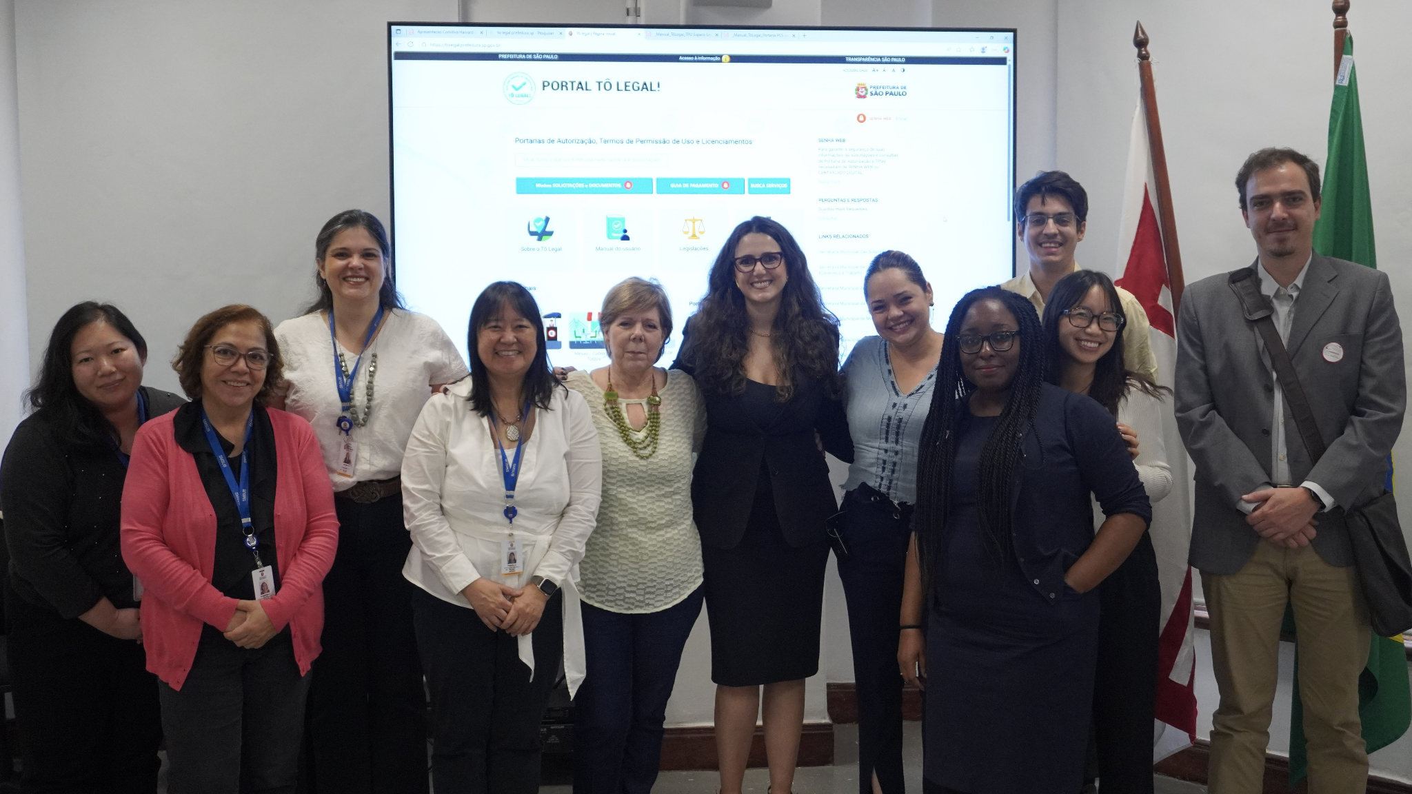 Fotografia de um grupo de dez pessoas, sendo sete mulheres e três homens, em frente a uma tela grande que exibe a página de um "Portal Tô Legal" da Prefeitura de São Paulo.  O grupo está posicionado lado a lado, sorrindo para a câmera. A maioria das mulheres veste blusas claras ou camisas sociais. O homem mais à direita veste um blazer cinza claro e calça cáqui, com uma pasta a tiracolo. O outro homem, mais ao centro-trás, veste uma camisa clara e sorri.  Ao fundo, à direita, há duas bandeiras, uma verde e outra marrom-escura, apoiadas na parede. A tela de projeção exibe a interface de um portal online com o cabeçalho "Portal Tô Legal!" e o logotipo da "Prefeitura de São Paulo". O ambiente é uma sala de reunião.