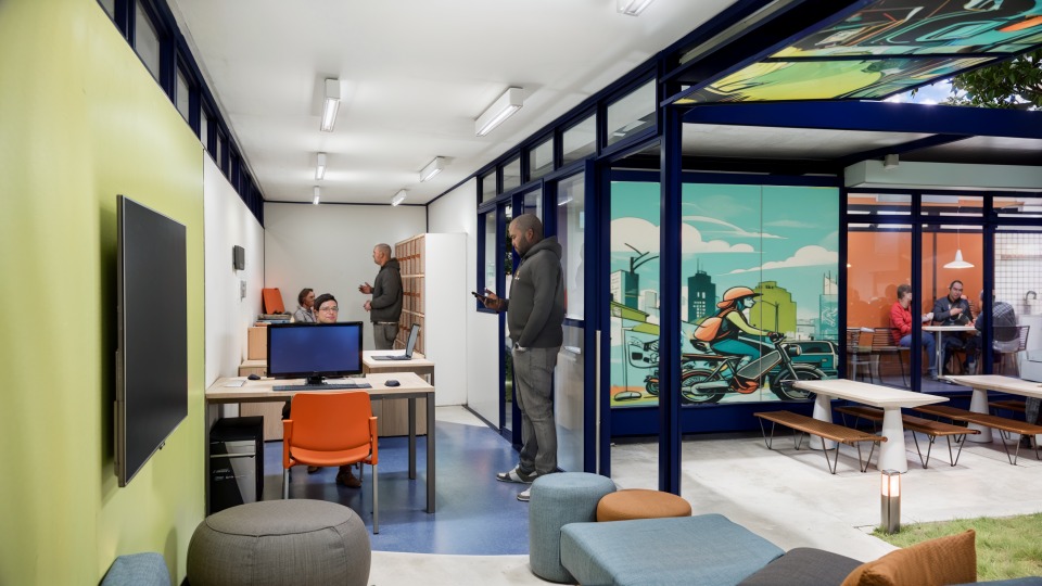 Fotografia de um espaço interno e externo moderno, bem iluminado e colorido, possivelmente um centro de apoio ou coworking.  O ambiente interno, à esquerda, tem paredes pintadas em branco e um tom de verde-limão. O piso é azul claro com detalhes em cinza. Há uma área de trabalho com mesas de escritório, um monitor de computador em destaque, e uma cadeira laranja. Algumas pessoas são vistas trabalhando nas mesas ao fundo e um homem está em pé ao centro, usando um celular.  O ambiente externo (ou uma área envidraçada) está à direita, separado por uma estrutura de esquadrias azuis-escuras. Esta área tem uma grande parede decorada com um mural vibrante em tons de azul-água, laranja e amarelo, que ilustra uma pessoa em uma motocicleta em meio a uma paisagem urbana. No canto inferior direito, vê-se um espaço de convivência com mesas e bancos, onde algumas pessoas estão sentadas, além de puffs coloridos e grama sintética ou natural, sugerindo uma transição para o ar livre.