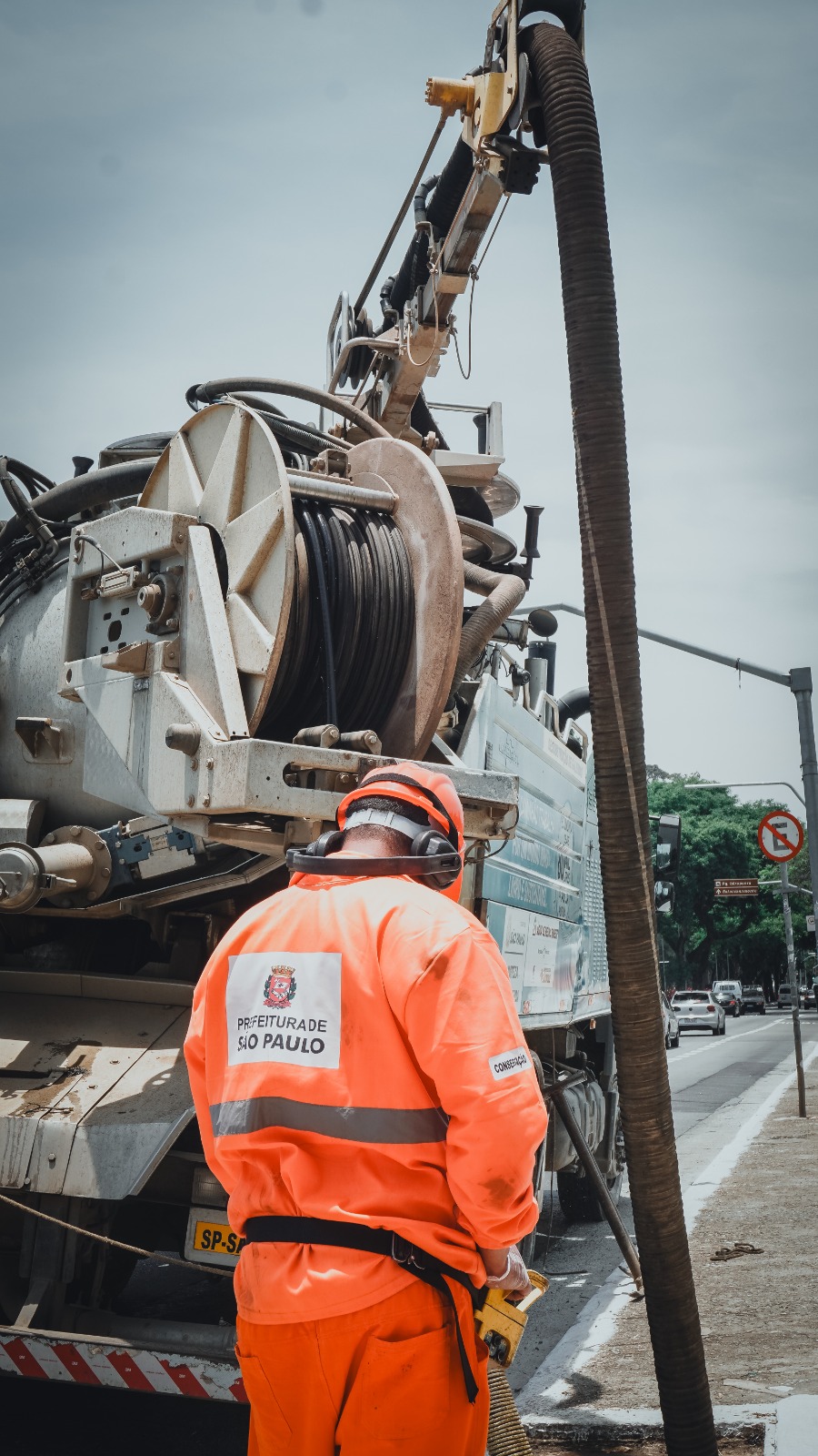 Trabalhador da Prefeitura de São Paulo, vestindo uniforme laranja com faixas refletivas e capacete com protetor auricular, atua na limpeza de rede subterrânea com caminhão hidrojato estacionado na via; o equipamento possui mangueiras grossas conectadas ao veículo, enquanto a operação ocorre em uma avenida arborizada, com carros passando ao fundo.
